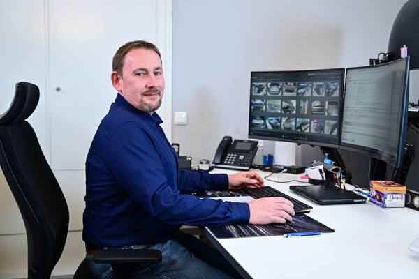 Mann im Büro am Schreibtisch mit zwei Monitoren und Fahrzeugfotos am PC