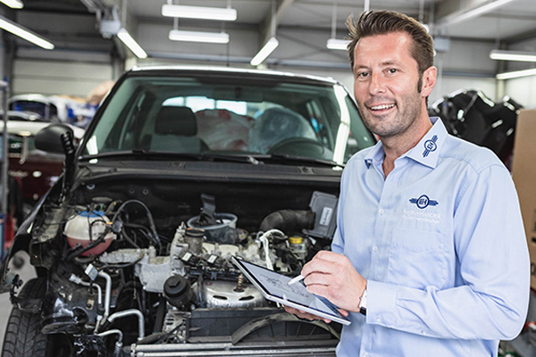 Ein Kfz-Gutachter prüft mit Tablet ein Auto mit offenem Motorraum in einer Werkstatt.